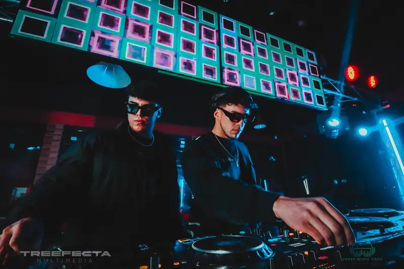 Dos DJs con lentes oscuros mezclando bajo una pantalla de luces